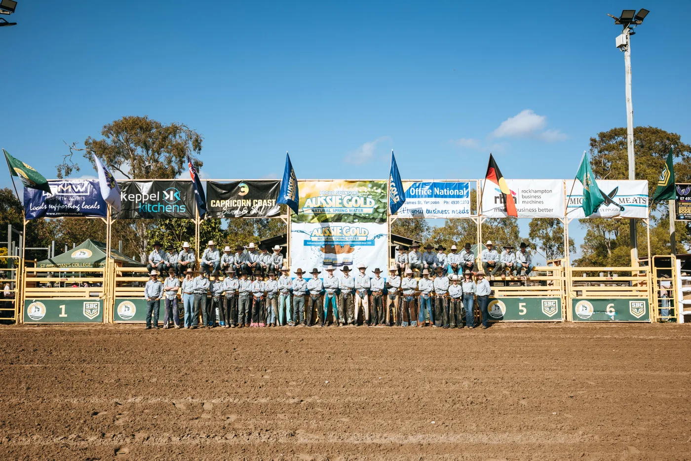 The full St Brendan's rodeo cohort lined up along the chutes in the school's own arena, sponsor banners and flags above them, under a clear Central Queensland sky.
