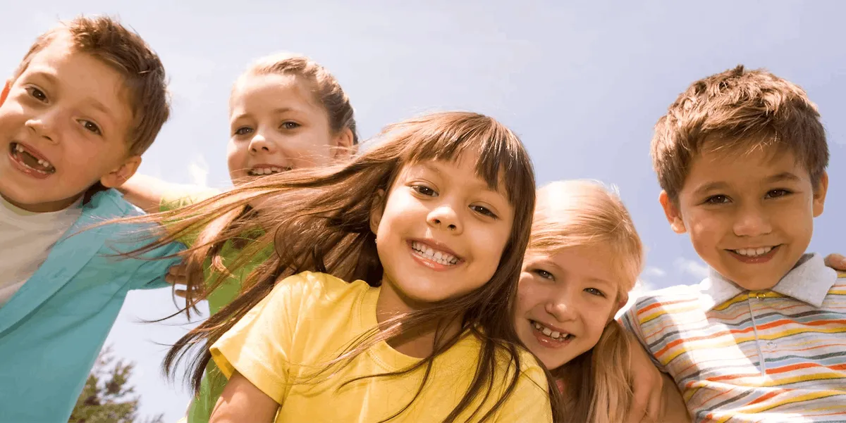 Five smiling primary school children huddled together outdoors under a blue sky